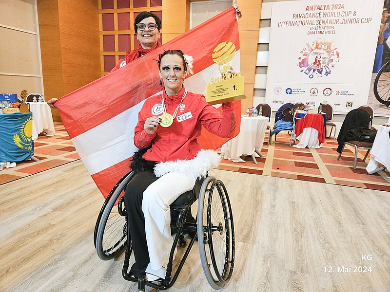 [Translate to Deutsch (Leichte Sprache):] Rollstuhl-Tänzerin Sanja Vukasinovic hält einen goldenen Pokal in der Hand. Ihre Trainerin Kerstin Govekar steht hinter ihr und spannt eine rot-weiß-rote Österreich-Flagge. Beide lachen in die Kamera. 
