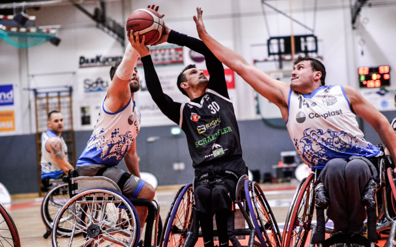 Ein Rollstuhl-Basketballer der FlinkStones hält den Ball in beiden Händen und setzt zum Wurf an. Zwei Spieler der Sitting Bulls greifen in Richtung Ball und versuchen ihn daran zu hindern.