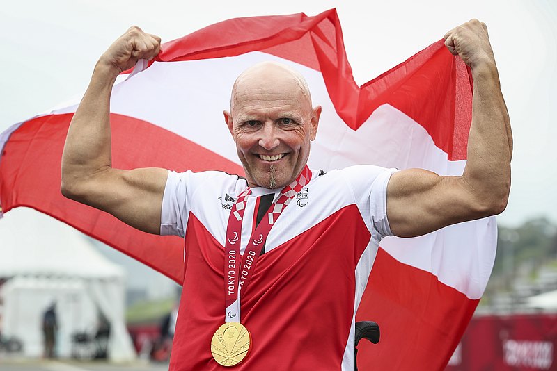 Walter Ablinger hält eine rot-weiß-rote Österreichfahne über den Kopf und lacht in die Kamera. Um seinen Hals hängt eine goldene Paralympics-Medaille. 