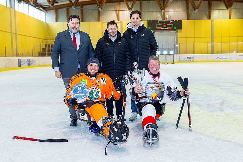 Para-Eishockeyspieler präsentieren am Eis ihre Pokale. Dahinter stehen die Trainer. 